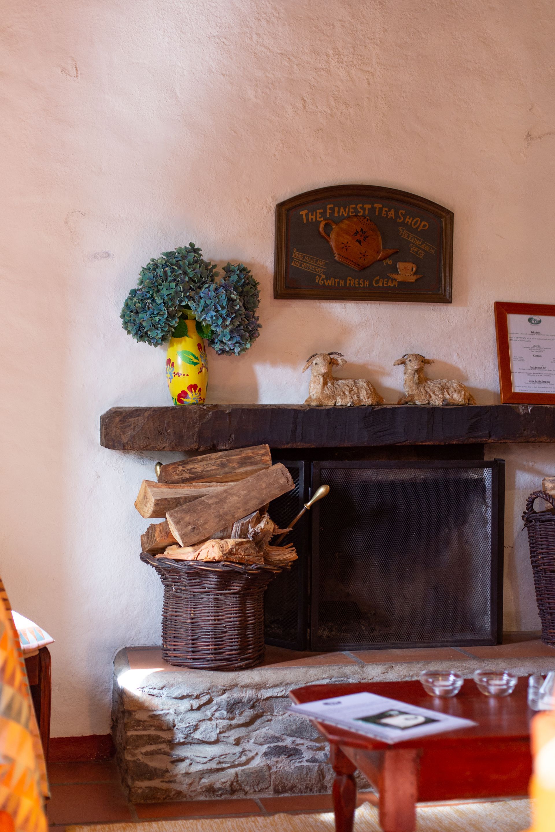 Fireplace with stacked logs, sheep figurines, flowers, and a sign in a cozy, rustic setting.