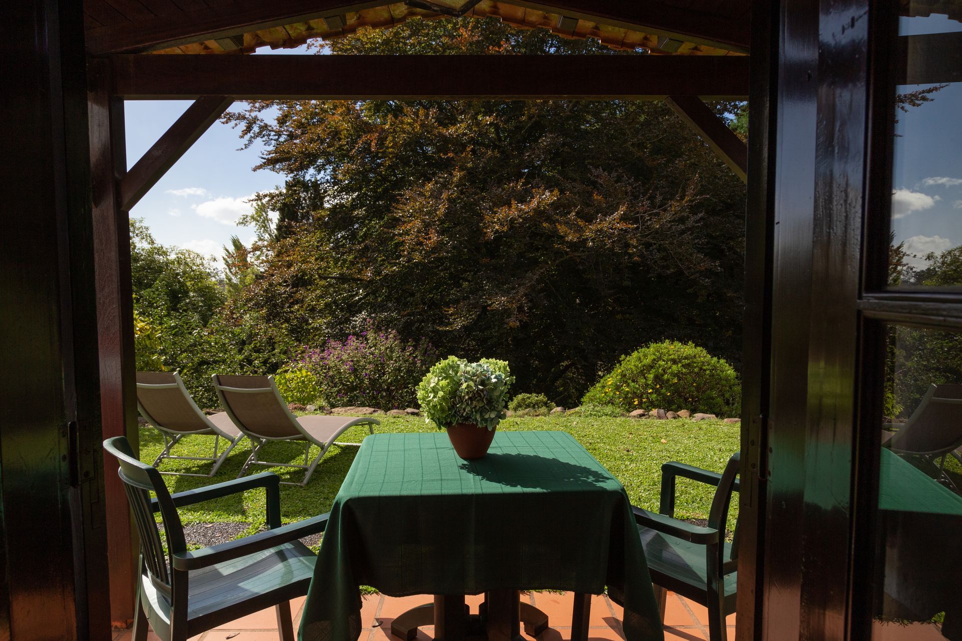 View from a wooden porch of a table set for two, facing a garden with lounge chairs and foliage.