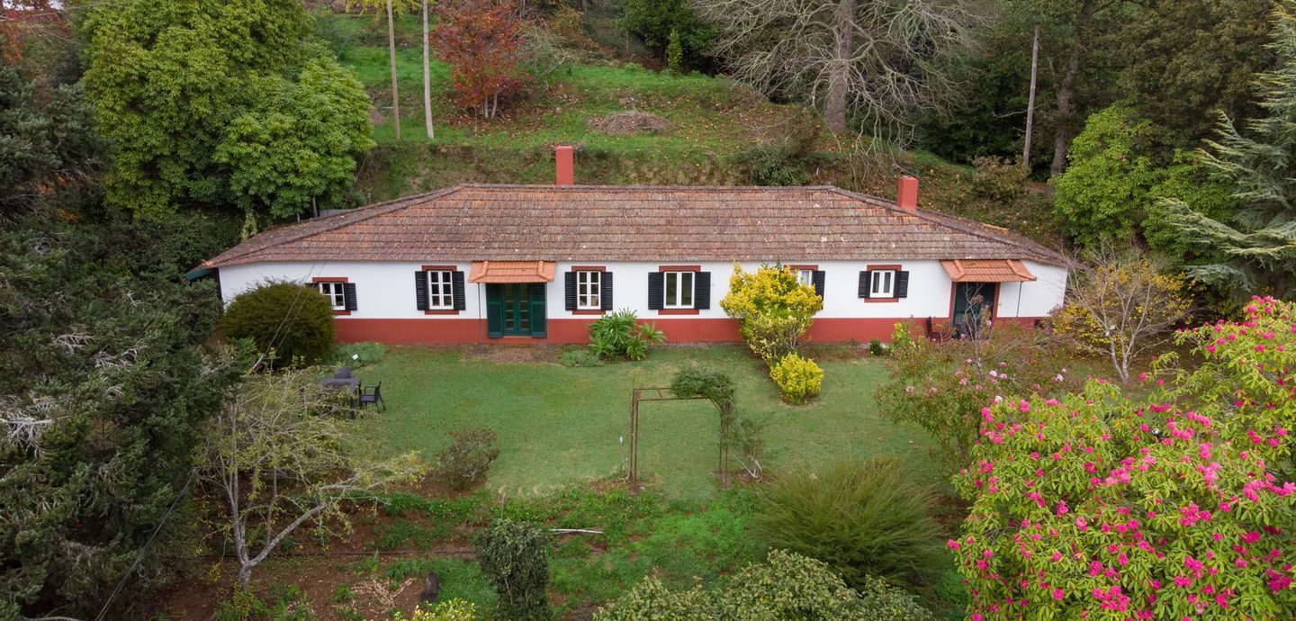 A white house with a brown tiled roof and green lawn, surrounded by lush trees.
