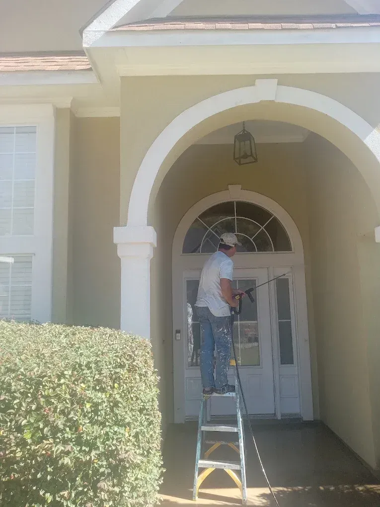 Man on ladder painting white arched doorway of beige house.