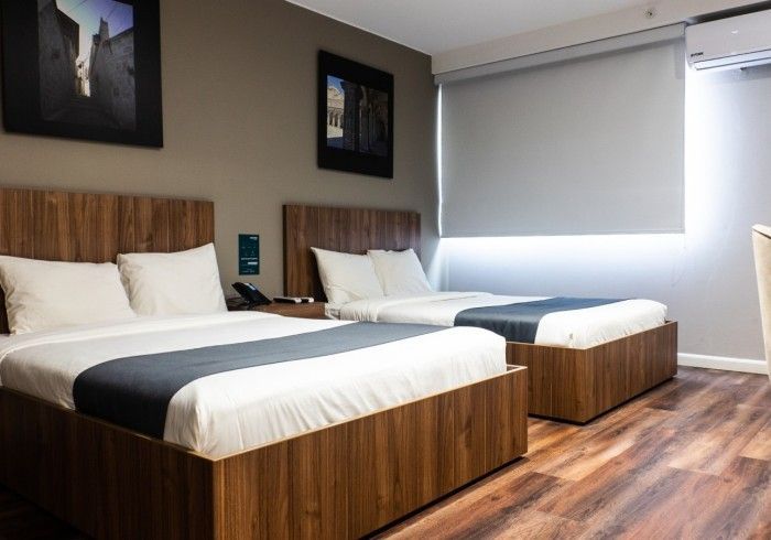 Dos camas en una habitación de hotel con detalles de madera, paredes grises y una ventana con persiana enrollable.