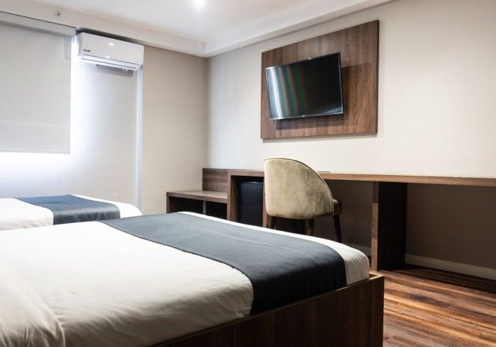 Habitación de hotel con dos camas, escritorio y televisor. Detalles en madera oscura, paredes neutras y luz natural.