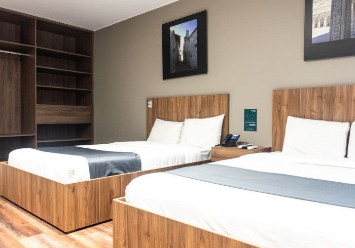 Habitación de hotel con dos camas, cabeceros de madera y armario. Paredes grises con cuadros.