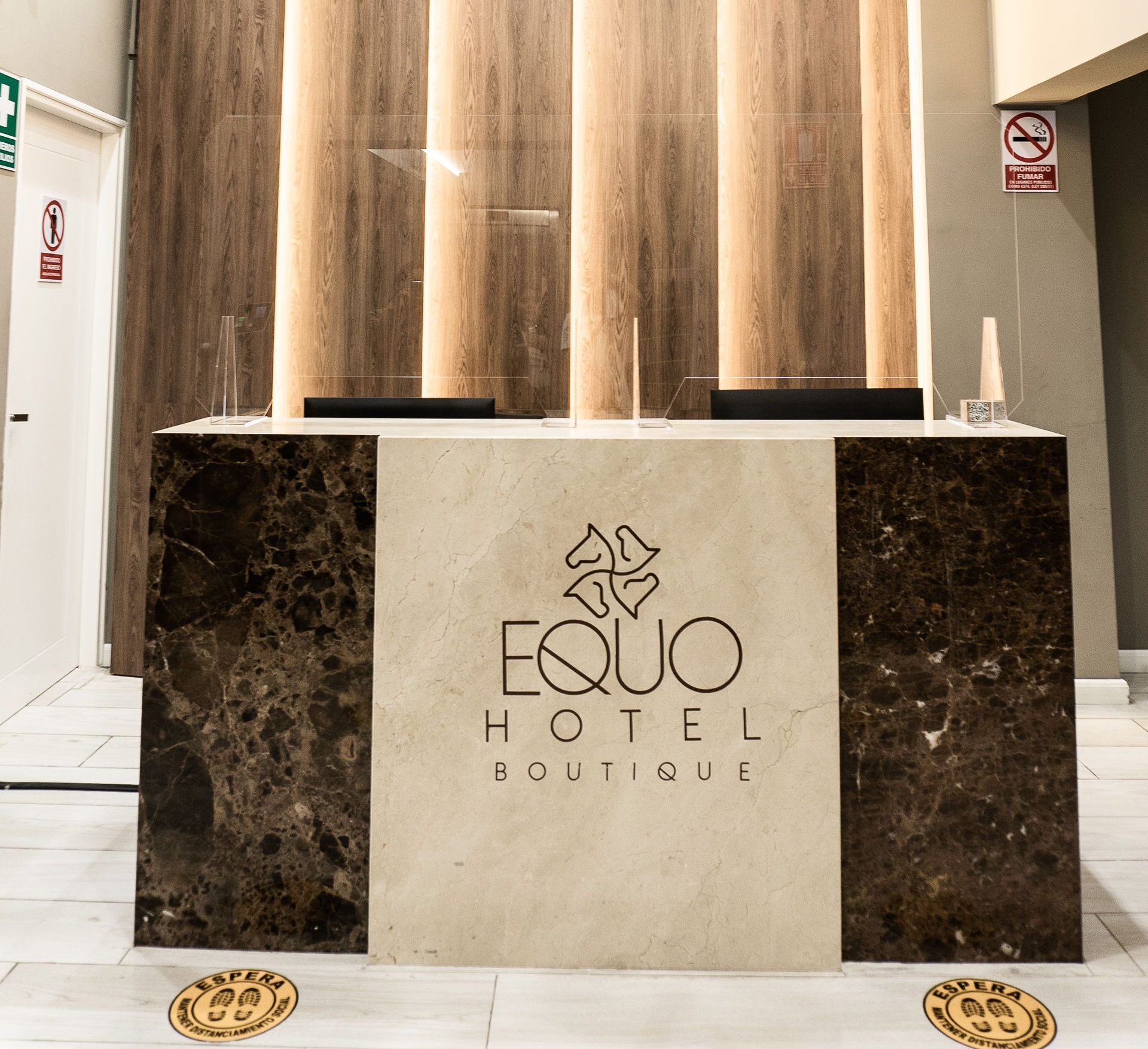 Mostrador de recepción de hotel con logo “EQÜO HOTEL BOUTIQUE”, detalles en mármol y barreras de seguridad.