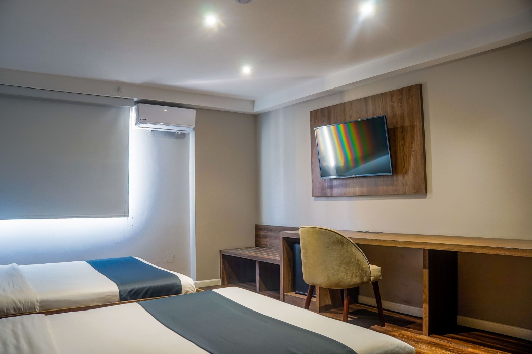 Dos camas en una habitación de hotel con detalles de madera, paredes grises y una ventana con persiana enrollable.