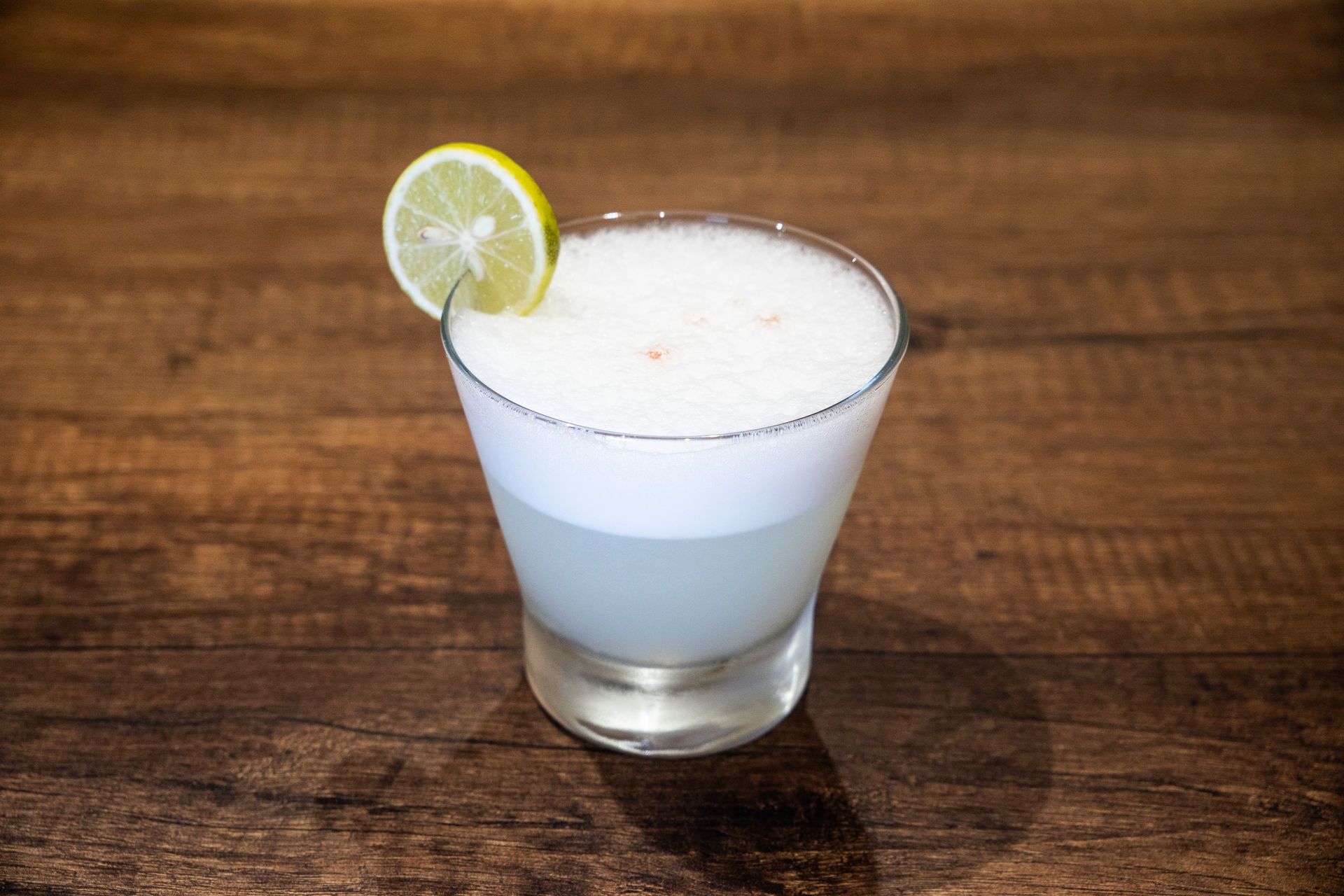 Cóctel en copa con cubierta blanca espumosa y guarnición de rodajas de lima sobre una mesa de madera.