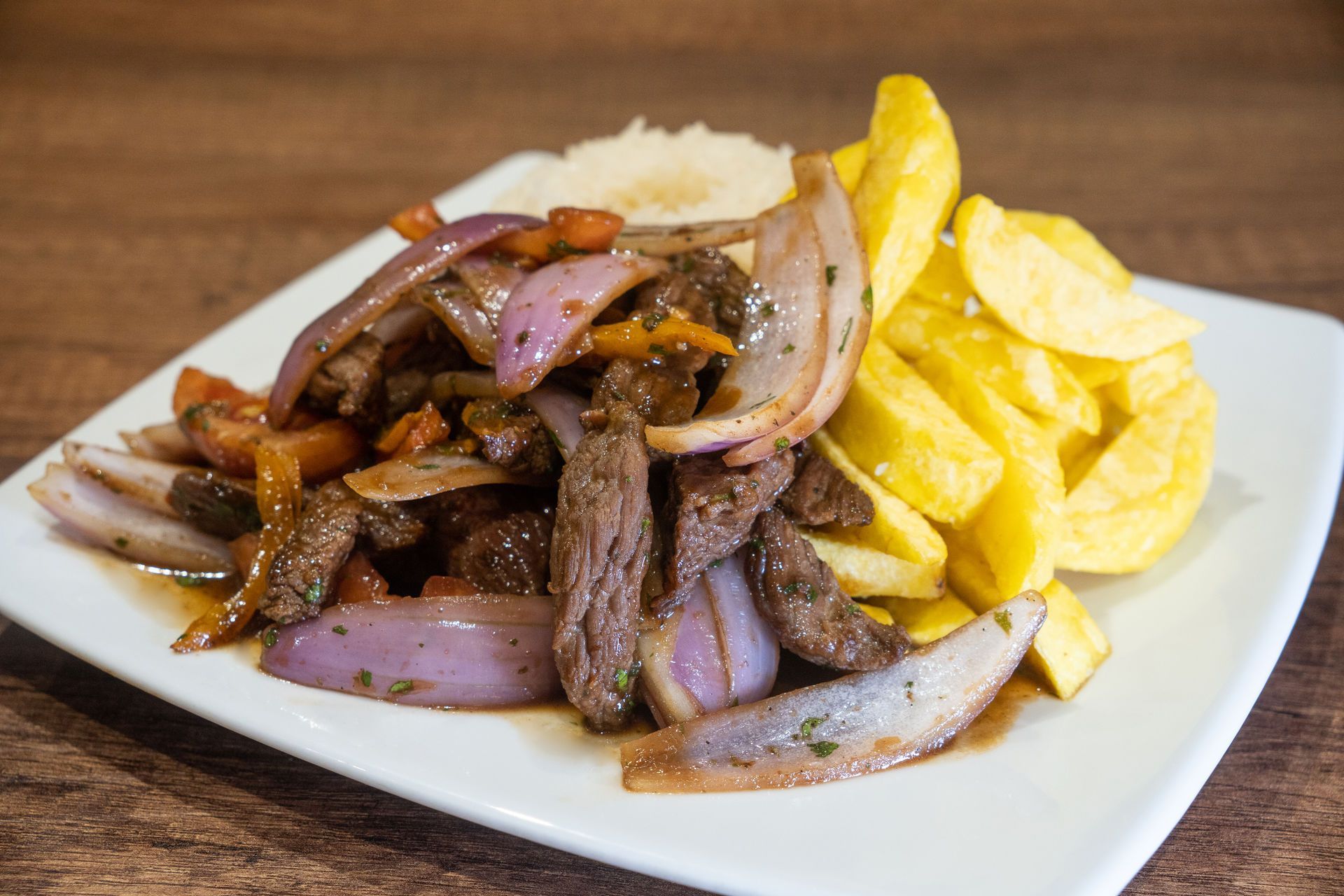 Lomo Saltado: Carne de res salteada con cebollas rojas, tomates y papas fritas, servida con arroz en un plato blanco.