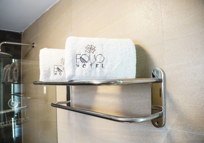 Dos toallas blancas enrolladas en un perchero metálico en el baño de un hotel. Llevan la etiqueta «EQUO HOTEL».