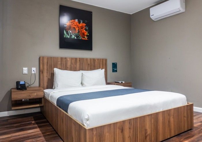 Dormitorio con marco de cama de madera, mesitas de noche, obras de arte y aire acondicionado en una pared gris.