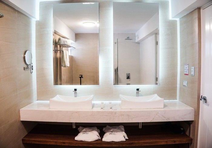 Baño con dos lavabos, espejos y un estante de madera debajo de la encimera. Toallas blancas en el estante.