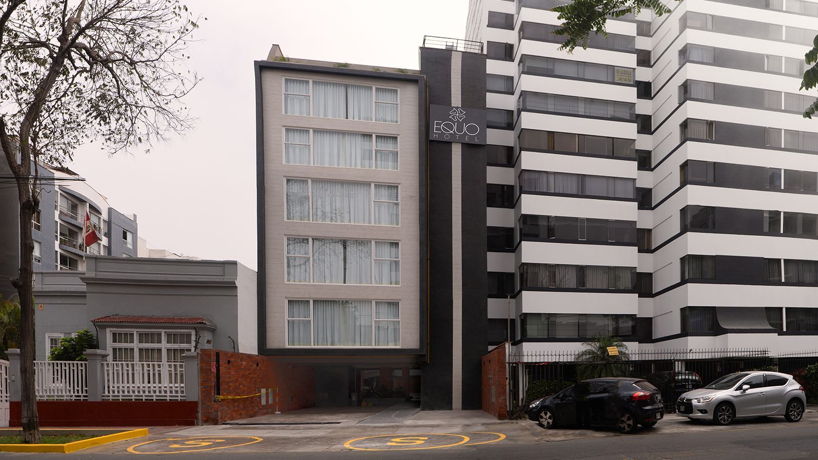 Edificio gris y blanco de varios pisos con reloj, autos estacionados en el frente, nublado.