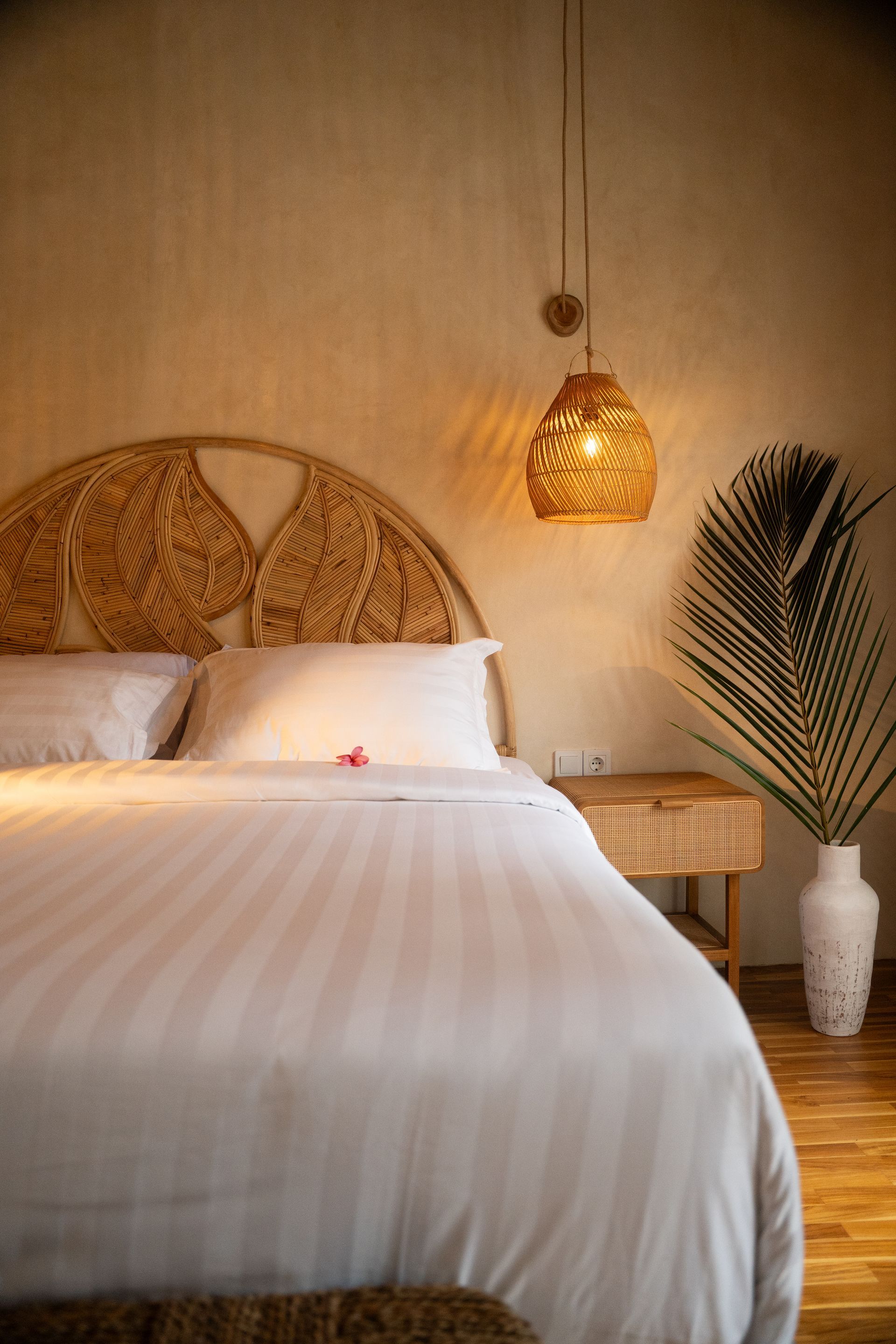 Bedroom with woven headboard and hanging lamp, white linens, and tall vase with greenery.