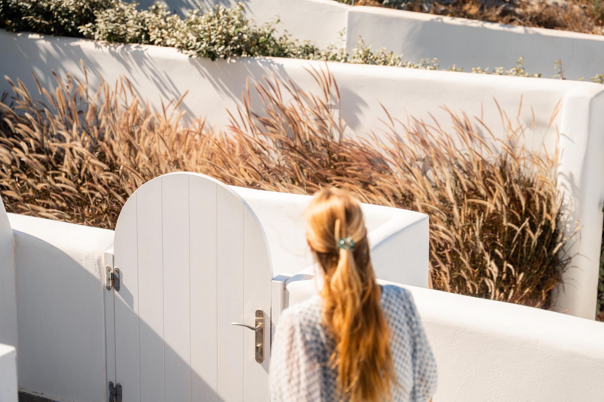 Woman with long blonde hair opens a white gate in front of tall brown grass, outdoors.