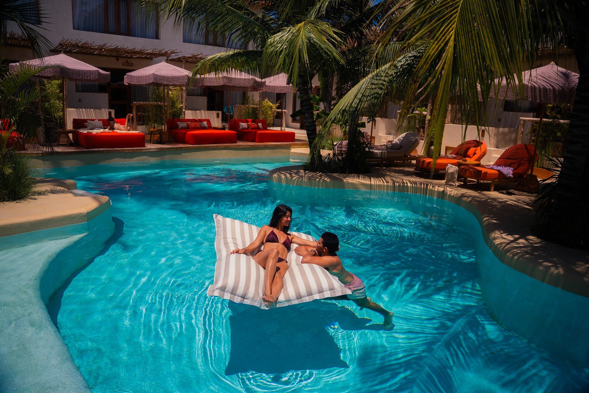 A couple relaxes on a large white and grey striped floating lounge chair in a bright blue swimming pool at a resort.