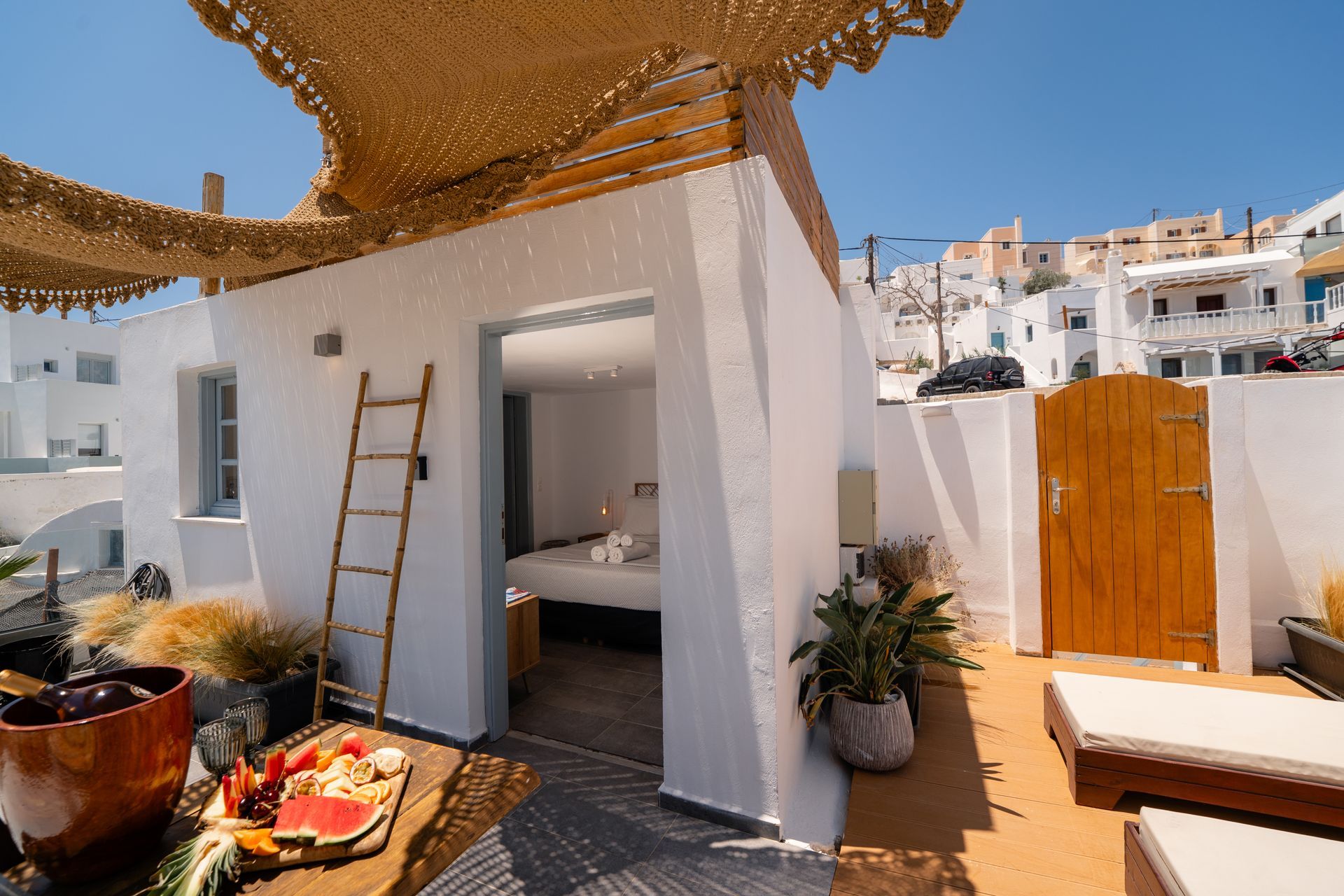 White-walled building with open doorway to a bedroom, shaded patio with lounge chairs, view of Santorini town.