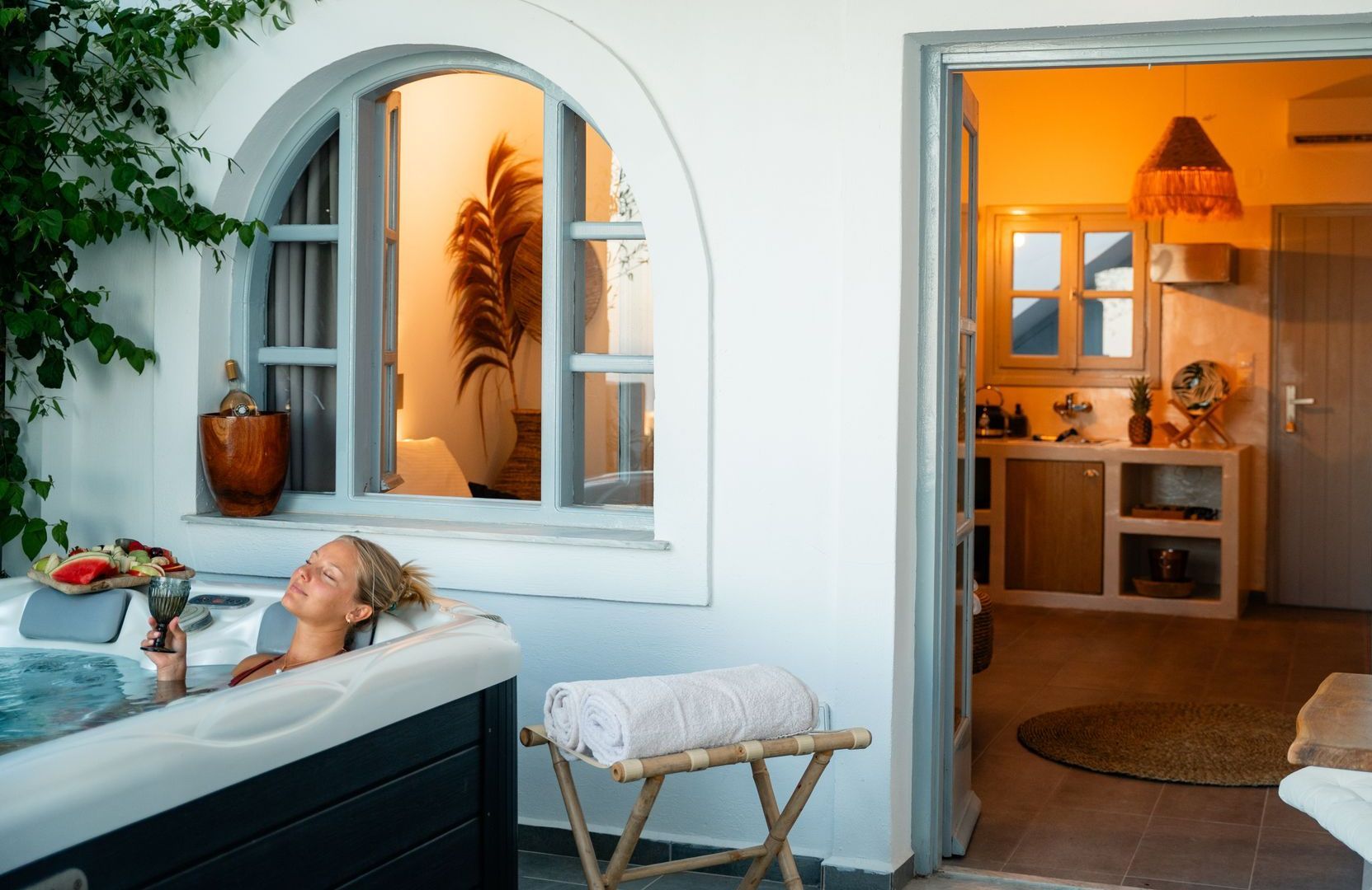 Woman in a hot tub with a drink, next to a window and an open doorway to a room with warm lighting.