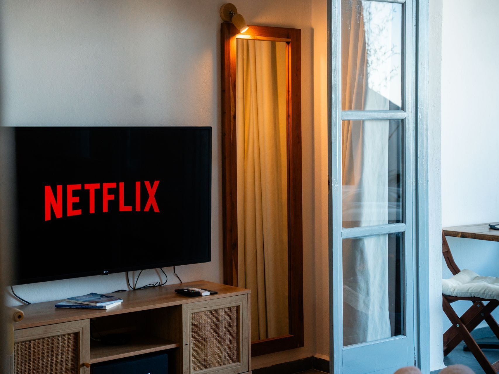 TV with Netflix logo on a wooden cabinet, next to a mirror and open door to a balcony with a chair.