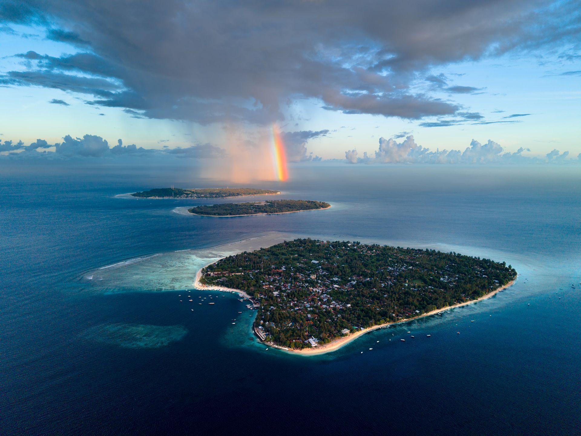 Aerial view of tropical islands in a deep blue ocean with a dramatic cloudy sky and a faint rainbow in the distance.