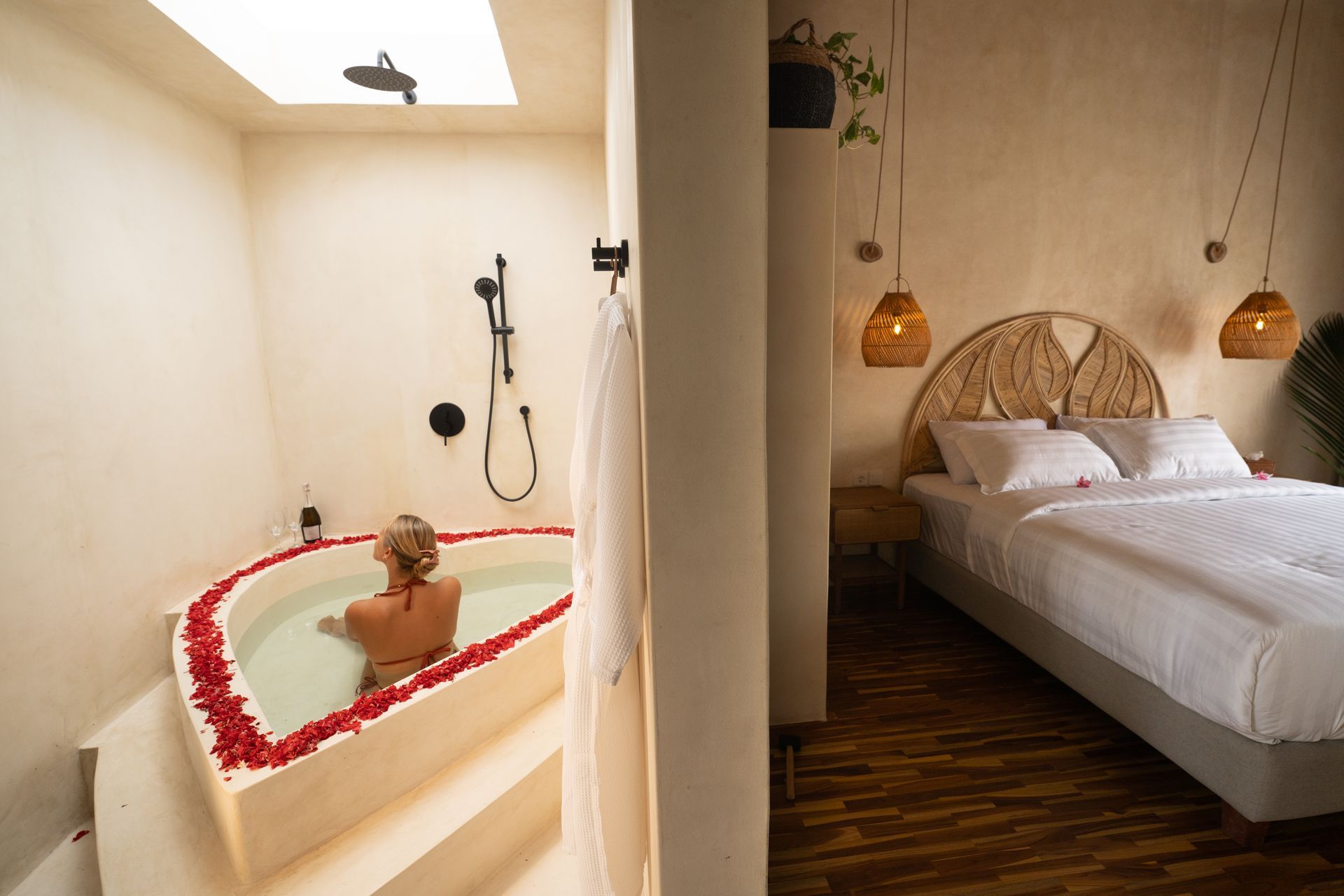 Woman in heart-shaped tub with rose petals; bedroom with bed, decorative lights. Light, airy room.