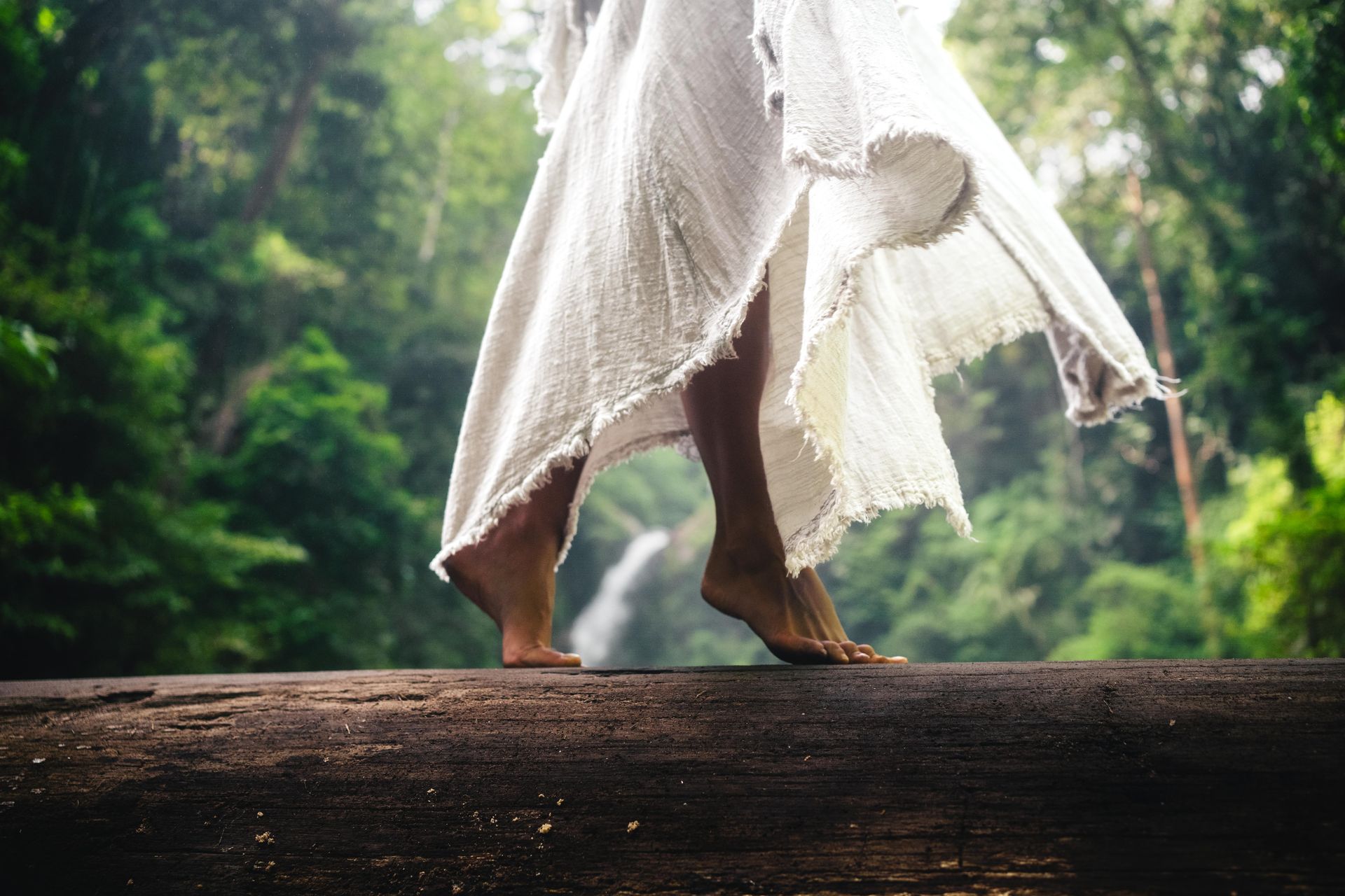A woman in a white dress is standing on a log in front of a waterfall.