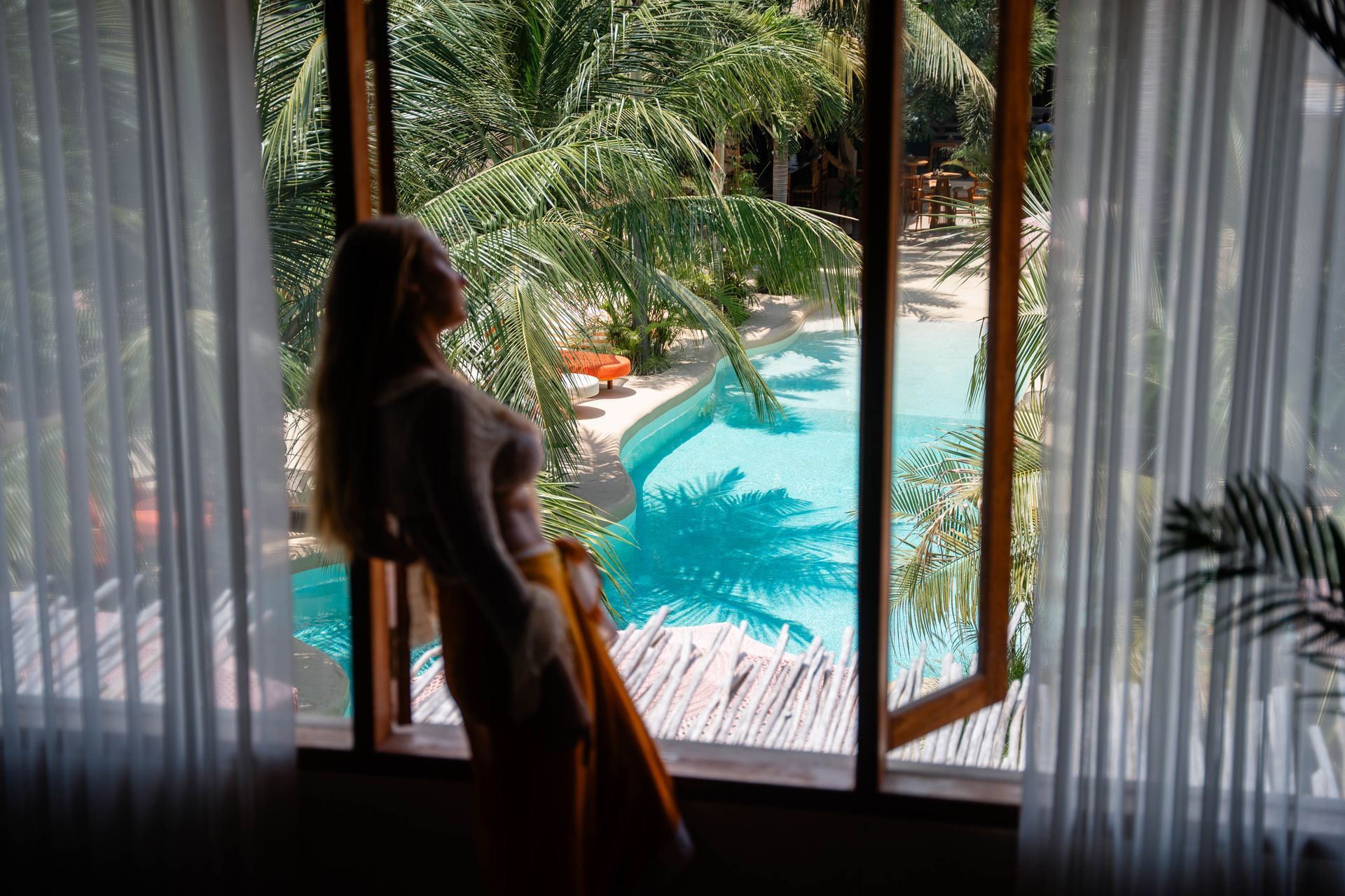 Woman leaning in a window, overlooking a pool surrounded by palm trees.