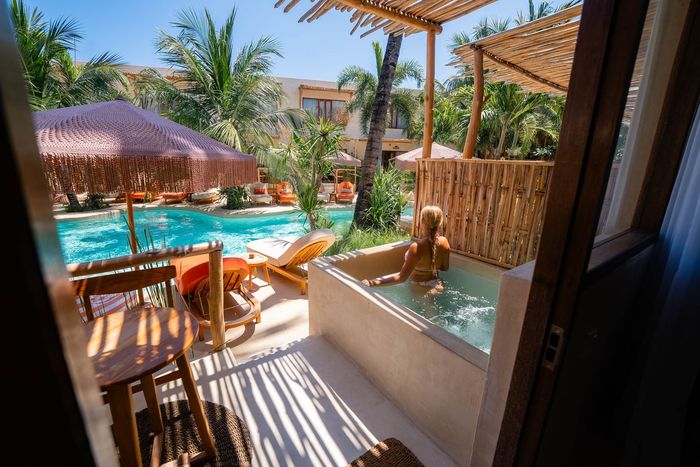 A woman in a swimsuit sits in an outdoor tub on a sunny patio, overlooking a pool.