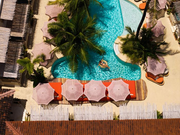 Overhead view of a turquoise pool surrounded by sunbeds with orange umbrellas, palm trees, and a person swimming.
