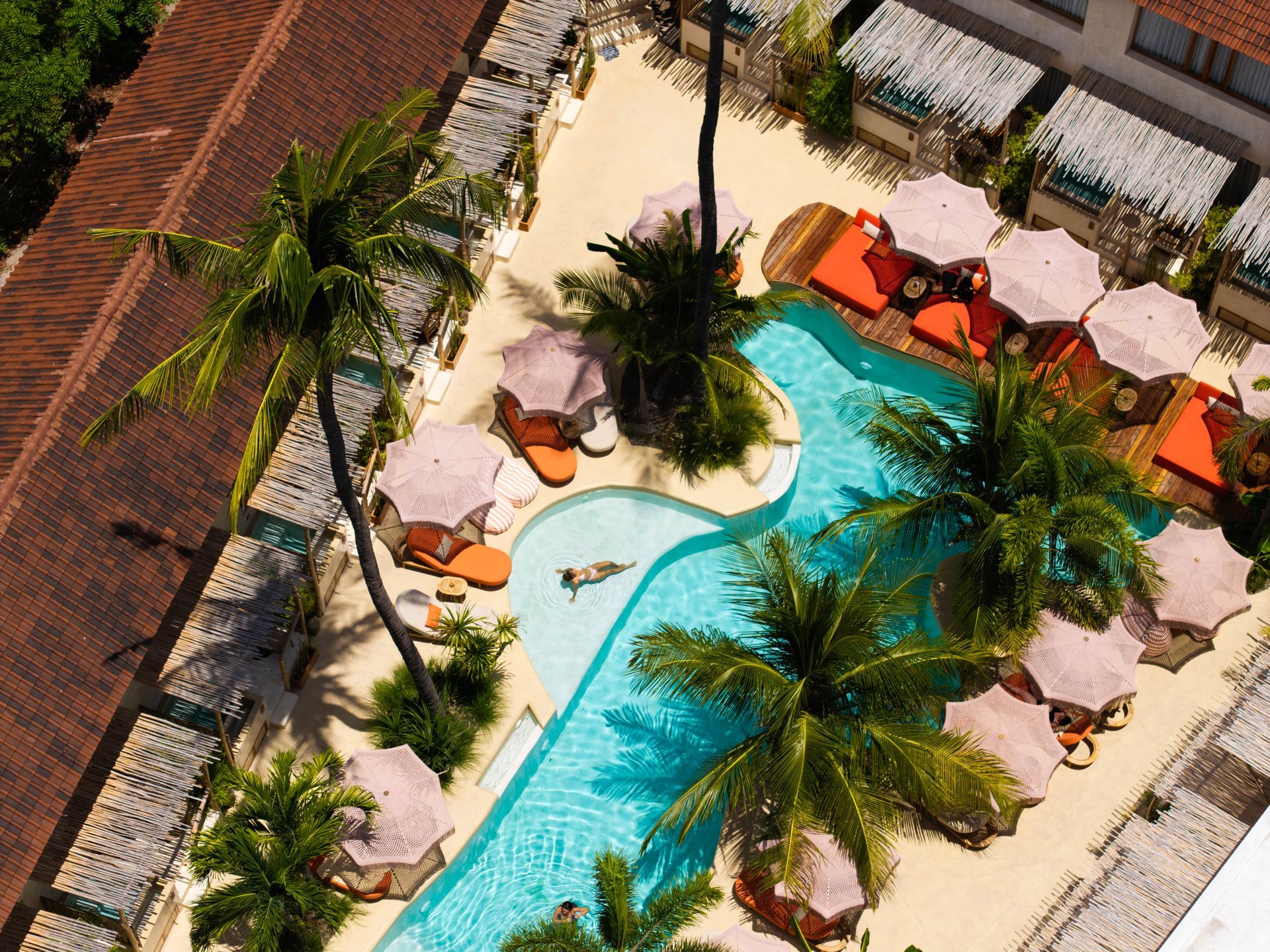 Aerial view of a pool area with a curvy pool, palm trees, umbrellas, and people sunbathing.