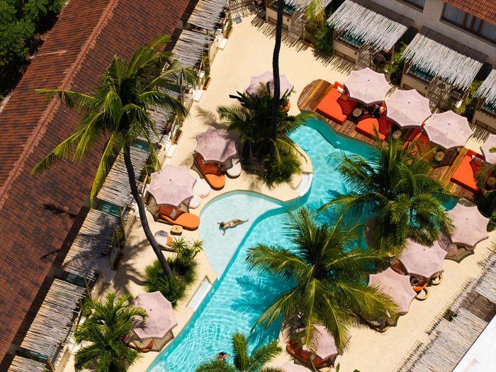 Aerial view of a pool area with a curvy pool, palm trees, umbrellas, and people sunbathing.