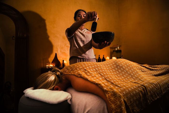 Woman receiving sound therapy at a spa. Therapist uses a singing bowl near the client. Dark, warm lighting.