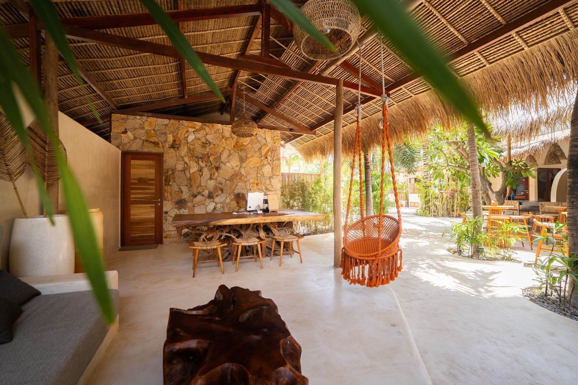 Open-air dining area with a stone wall, wooden table, hanging swing, and straw roof.