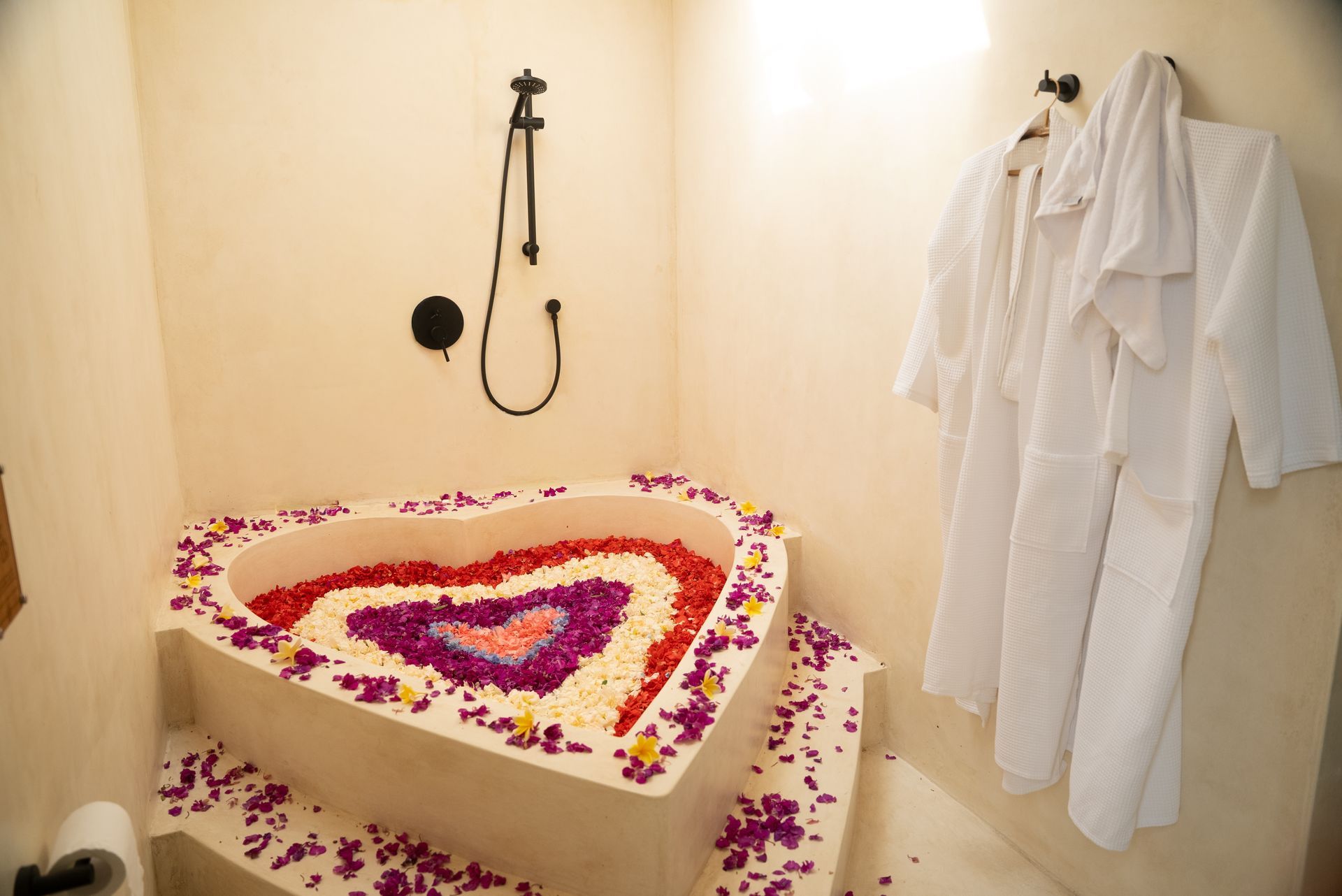 Heart-shaped bathtub filled with flower petals, next to a shower and white bathrobes hanging on the wall.