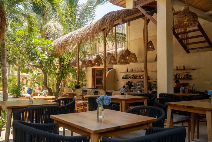 Outdoor restaurant with wooden tables and woven chairs under a thatched roof, surrounded by greenery.