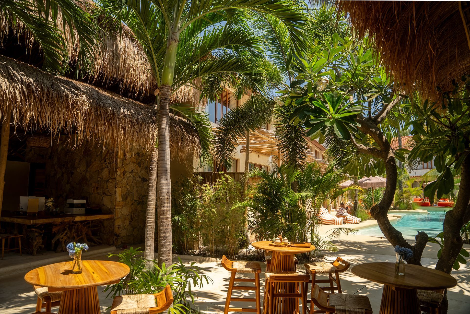 Outdoor dining area with thatched roof, wooden tables and chairs, tropical plants, and a pool.