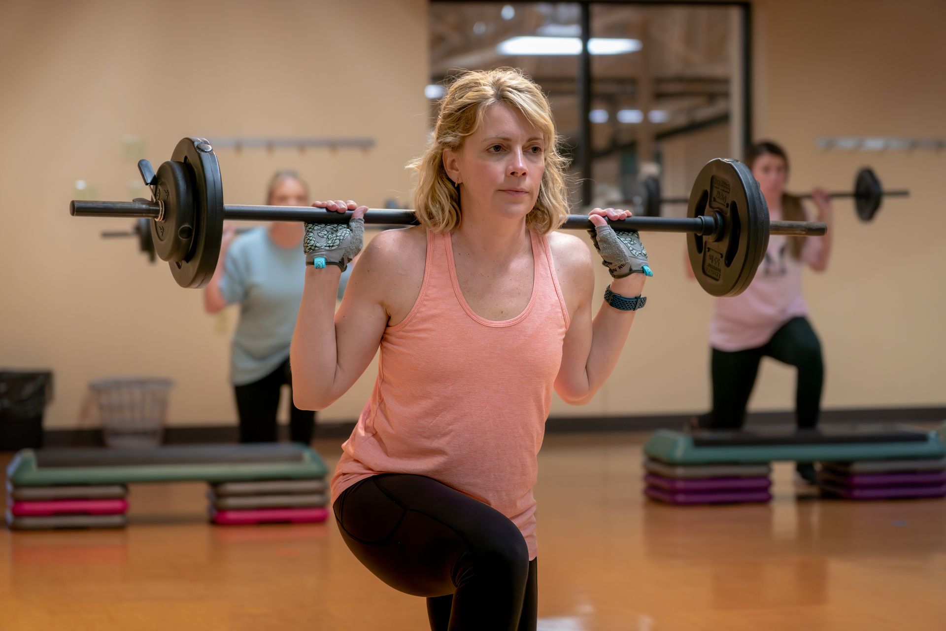 Woman doing barbell lunges in a gym, wearing pink tank top, black leggings, other women in background.