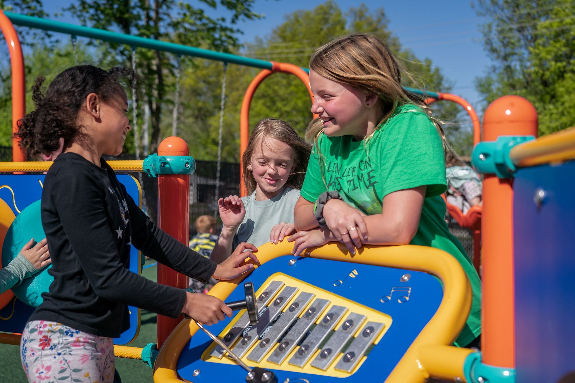 Three girls smiling and playing an outdoor musical instrument on a playground.