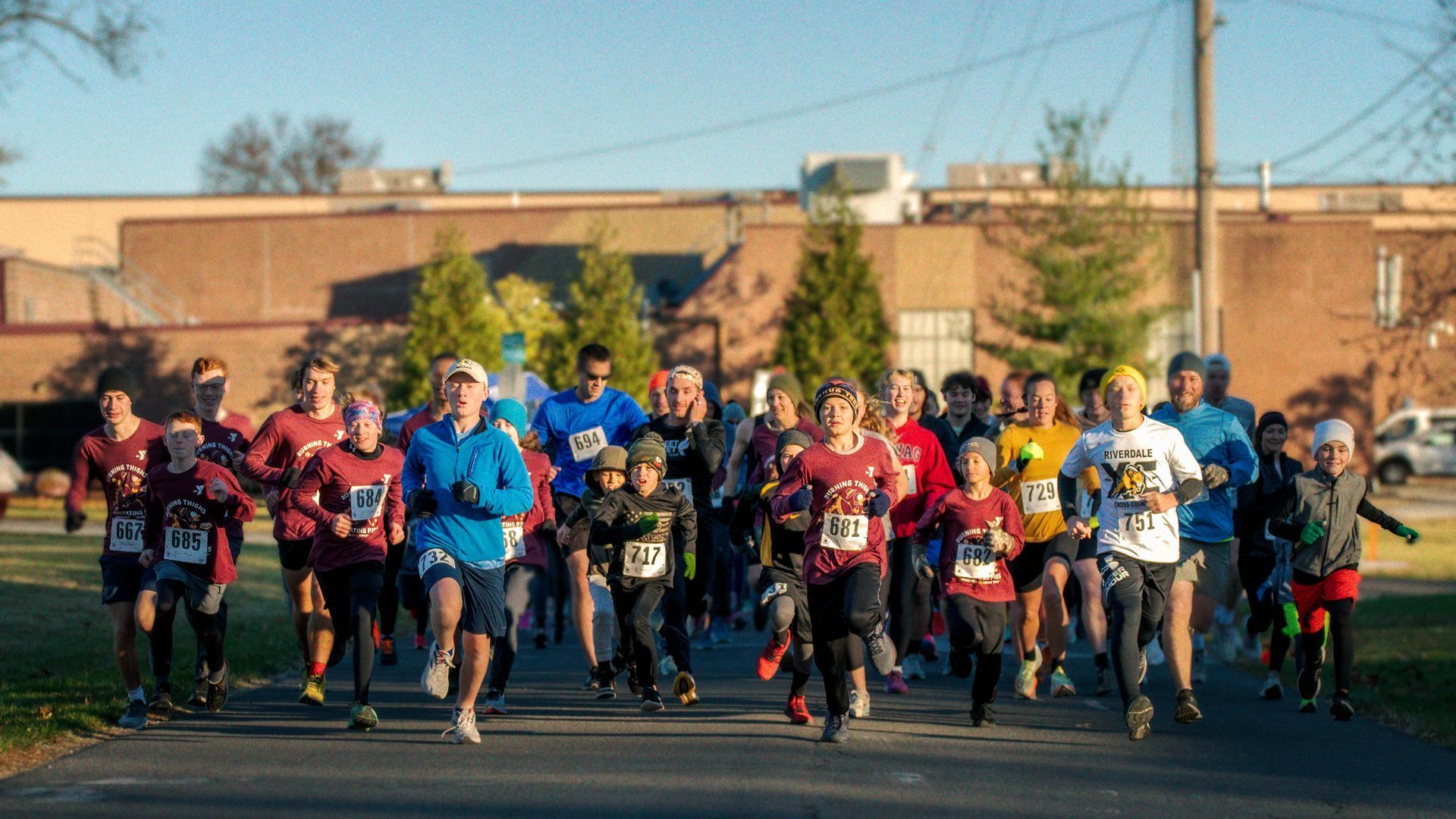 Runners in a race, front view. Many wearing running gear, starting line, sunny day, school building in background.