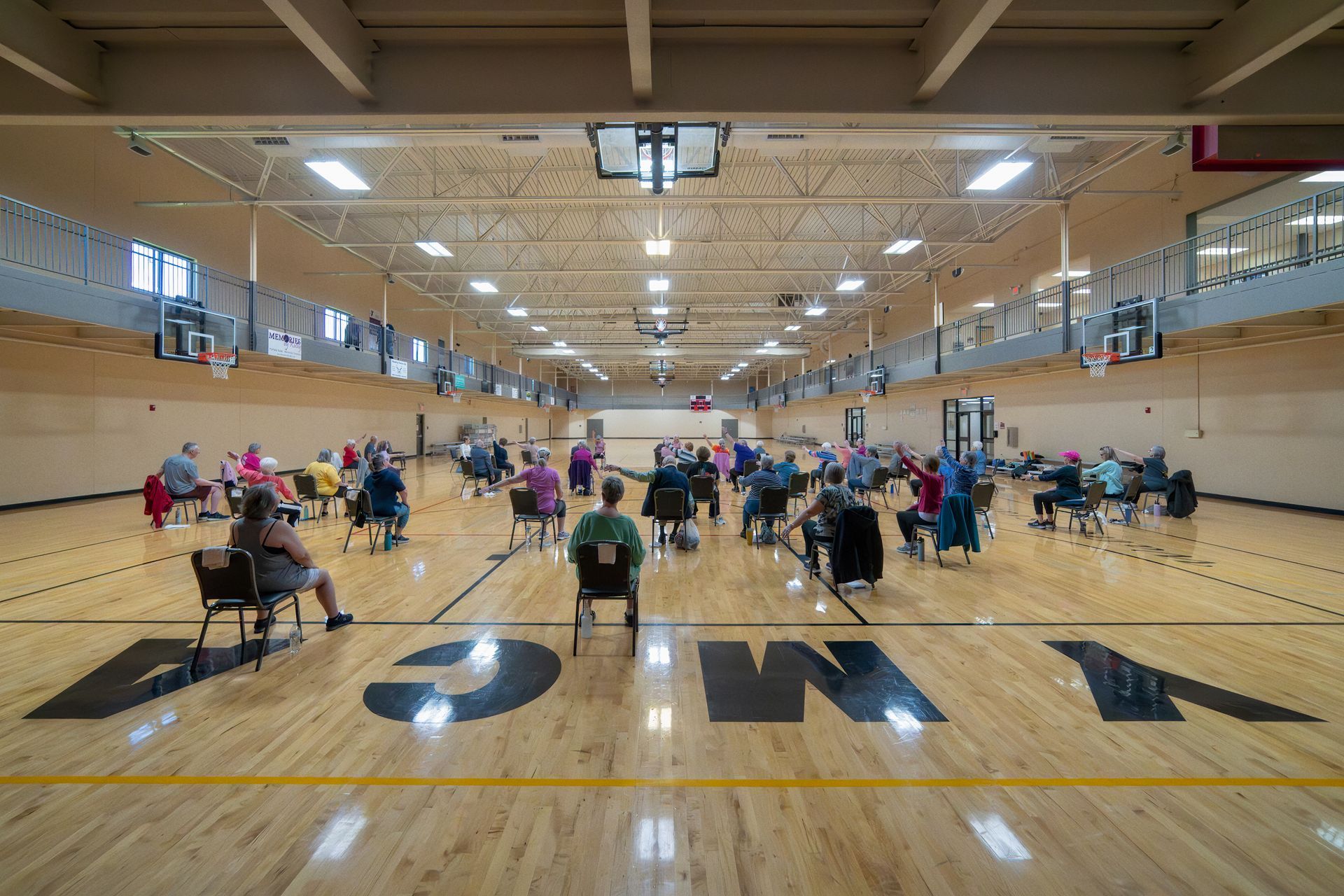 People seated in chairs on a gymnasium floor, likely attending a class. YMCA logo visible.