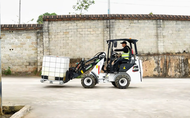 Small loader with a driver carrying white containers near a brick wall.