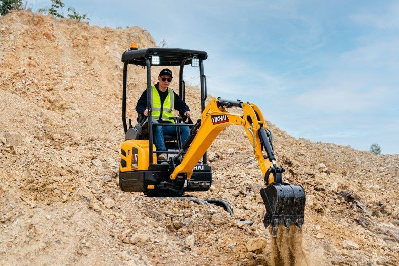 A person operating a yellow YUCHAI U17 excavator on a hillside, wearing a safety vest and sunglasses.