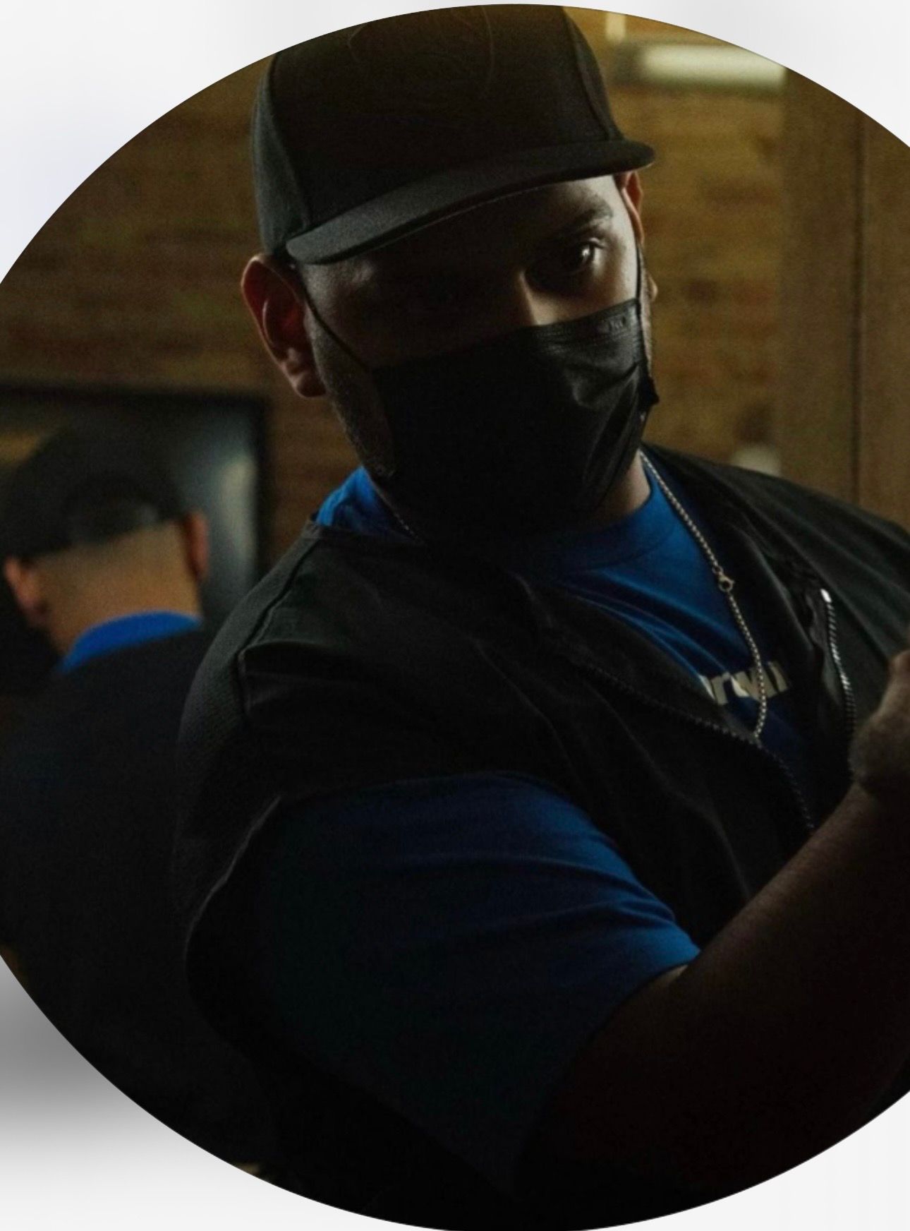 A person wearing a black cap, face mask, and blue shirt stands in a workspace, reflected in a mirror behind them.