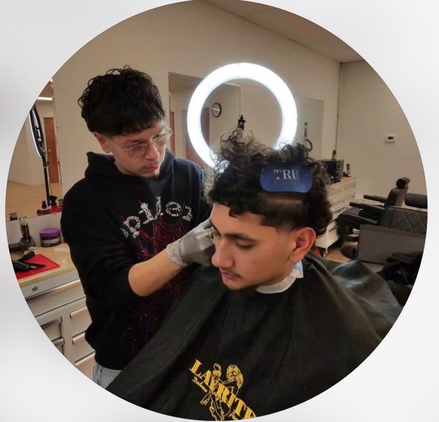 A barber wearing a black hoodie cuts a client's hair in a brightly lit salon with a circular ring light in the background.