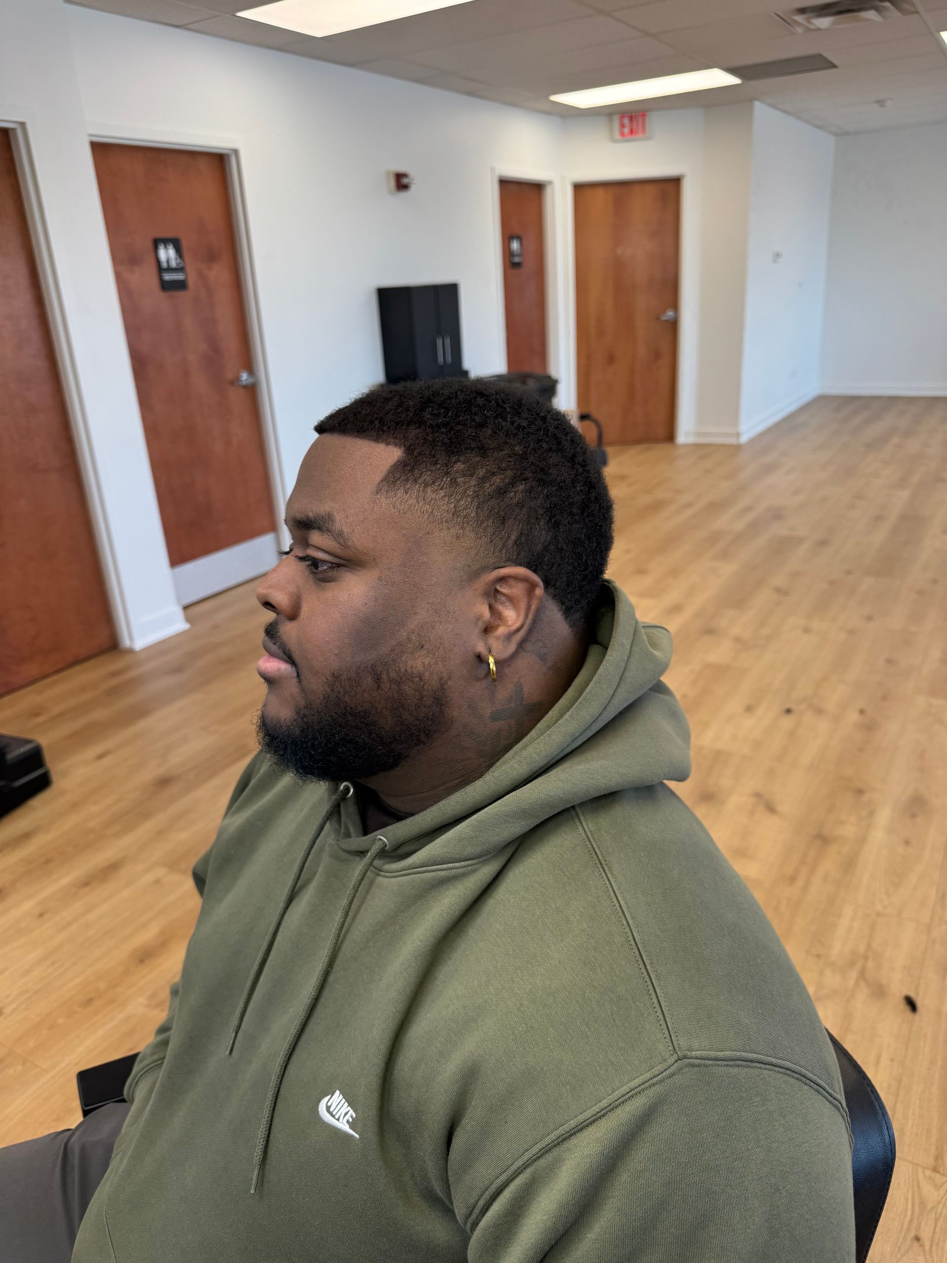 A person with a fresh haircut and a beard sitting in a barbershop wearing an olive green hoodie.