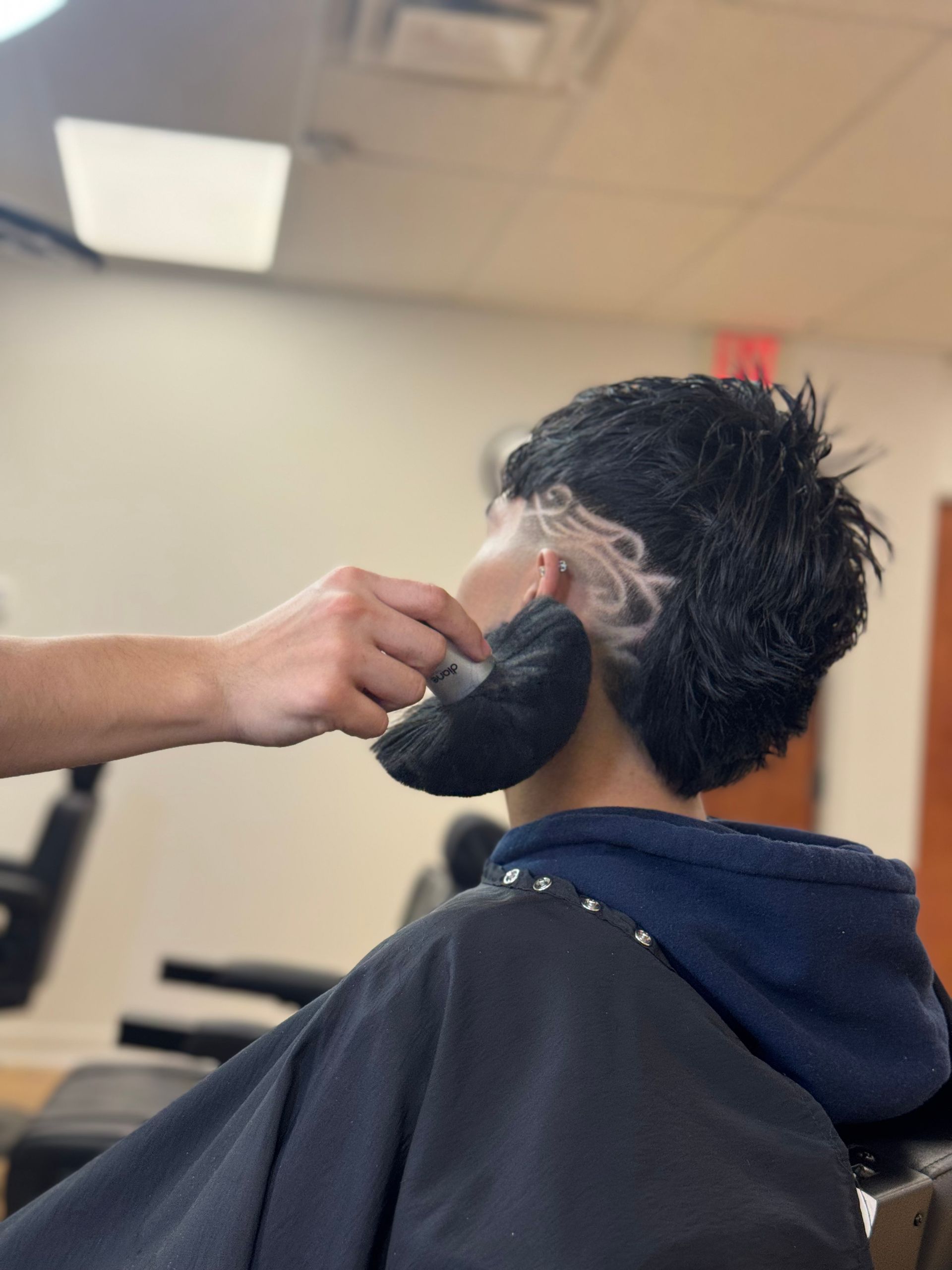 A barber brushes a client's neck with a black neck duster, highlighting a detailed, carved-in hair design on the side.
