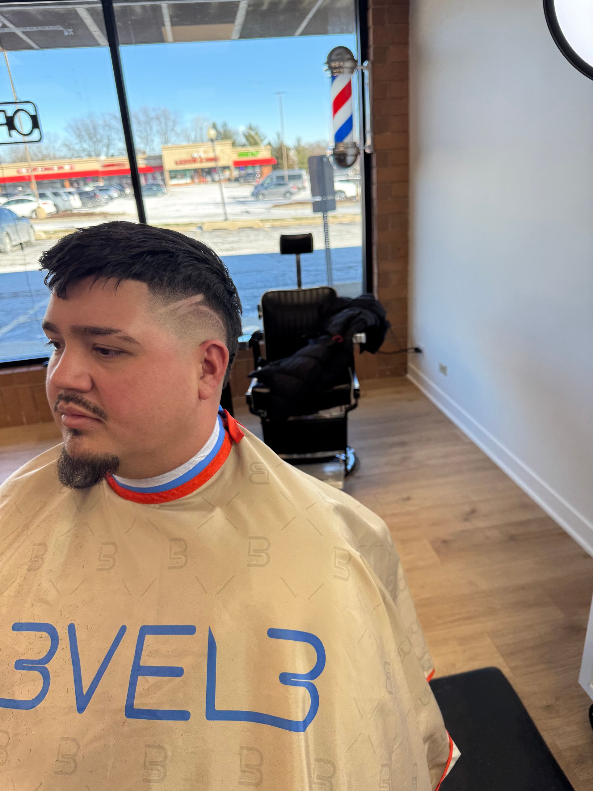 A person in a barber cape with a fresh haircut featuring a fade and a side part line, seated in a barbershop.