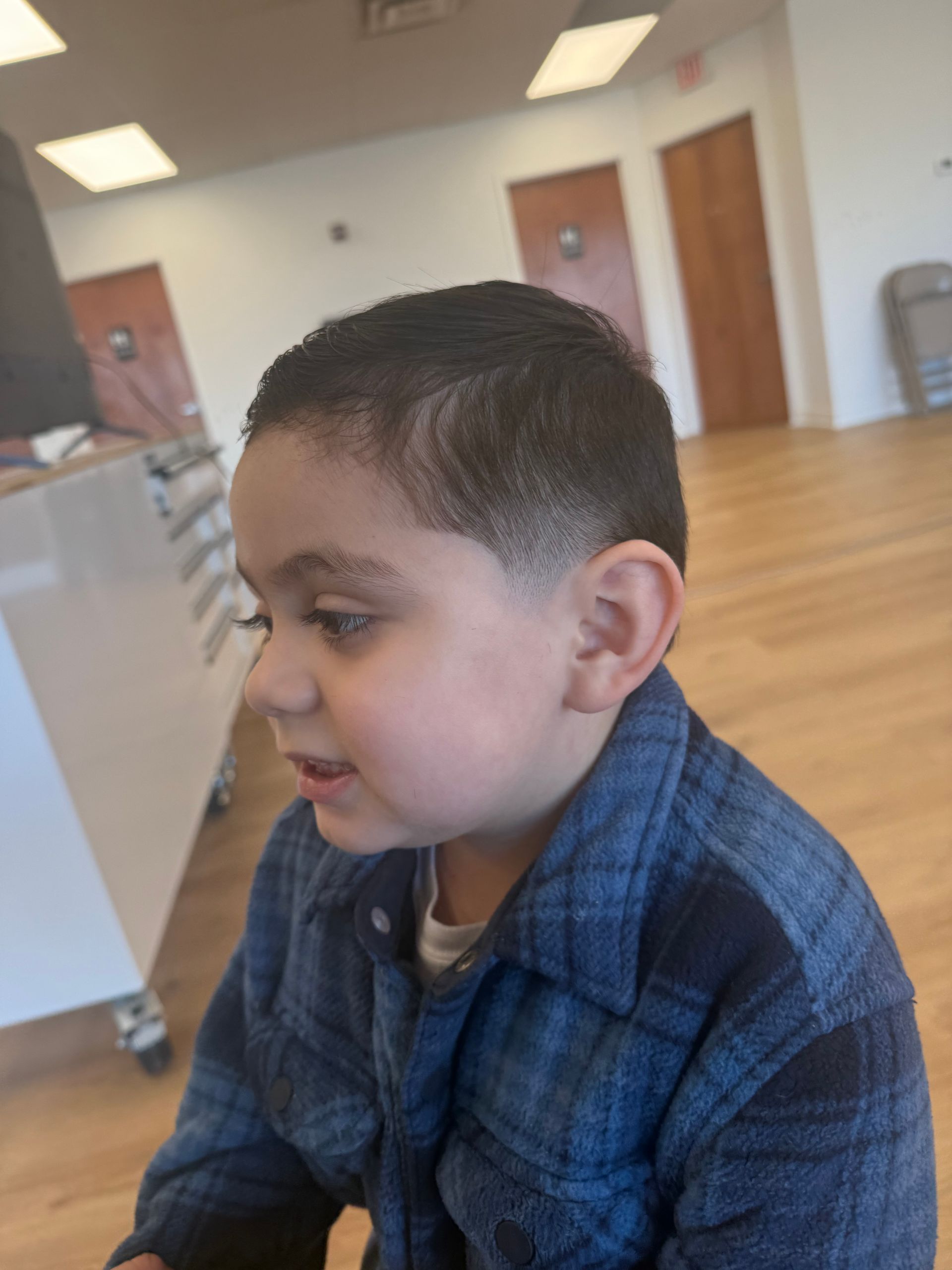 A child with a neat, short fade haircut and a blue plaid button-up shirt sits indoors, looking to the left.