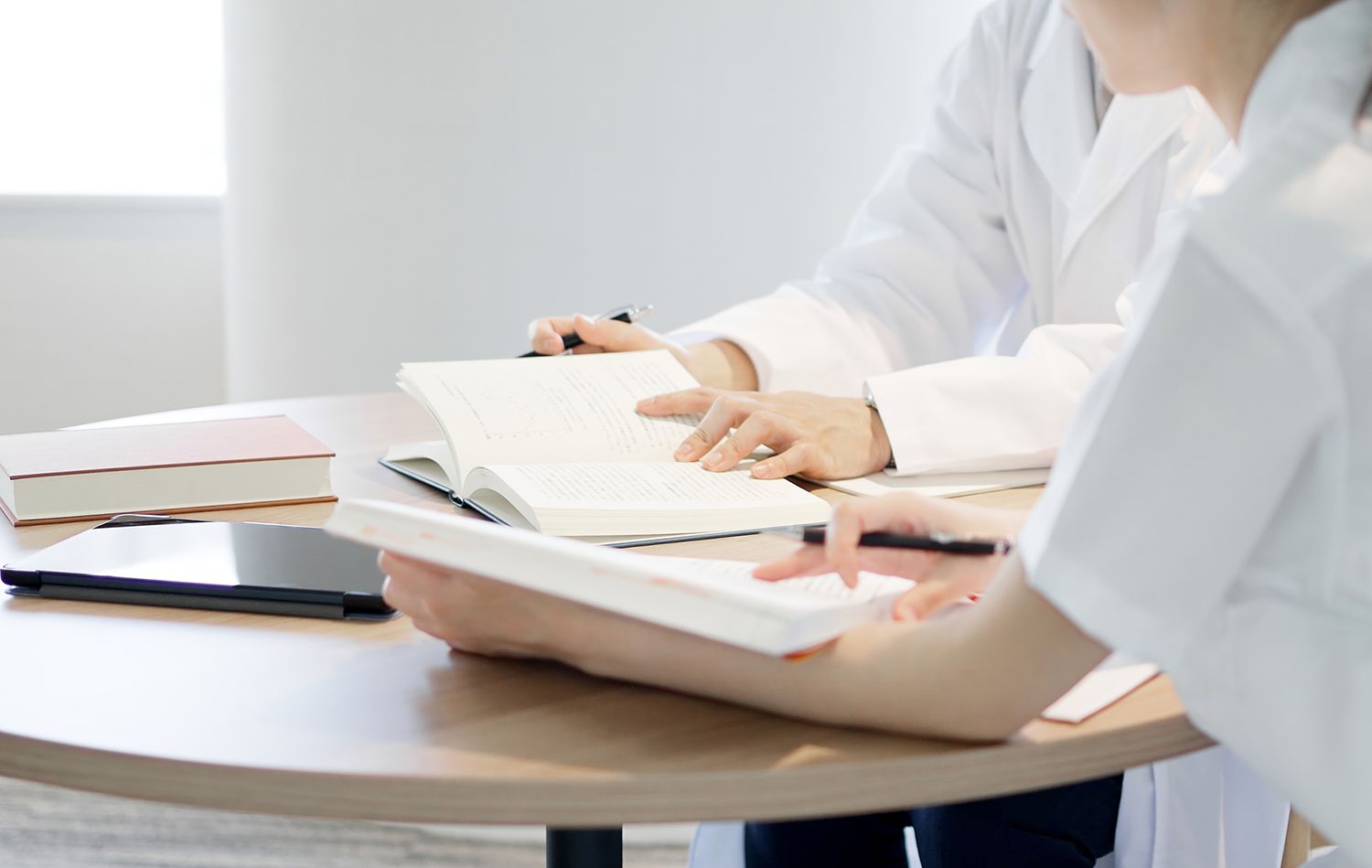 Two people in white coats study books at a table, likely medical professionals reviewing information.