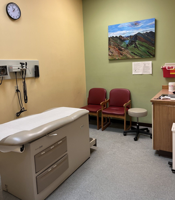 An exam room with a medical table, two red chairs, a rolling stool, a wall clock, and a mountain landscape painting.