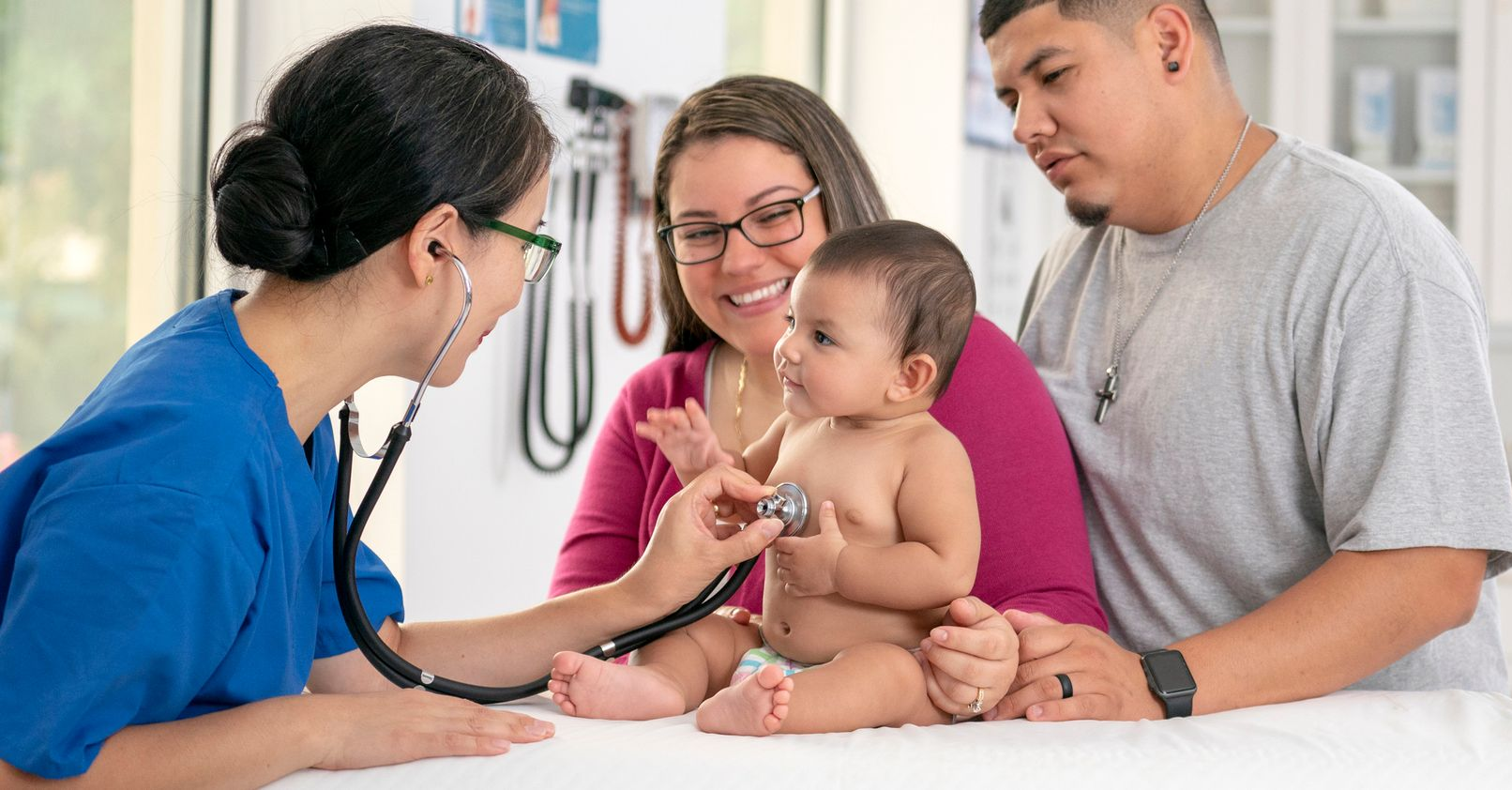 Doctor examines baby with stethoscope, parents watching at clinic.