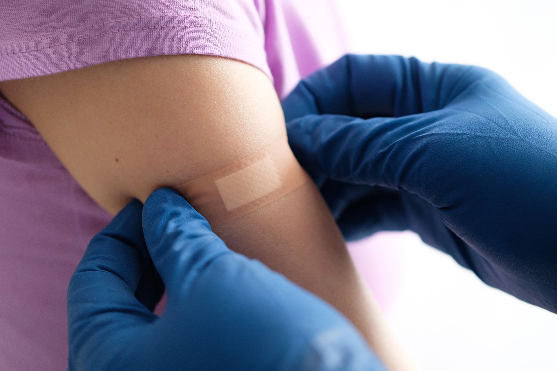 Blue-gloved hands applying a bandage to a child's arm after a vaccination, against a purple shirt.